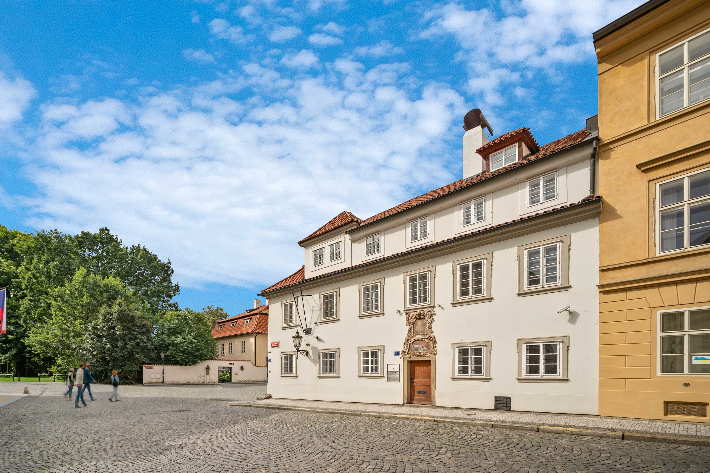 The Blue Fox House by day from Na Kampě square: white facade, red roof, the Baroque portal, the trees of Kampa on the left