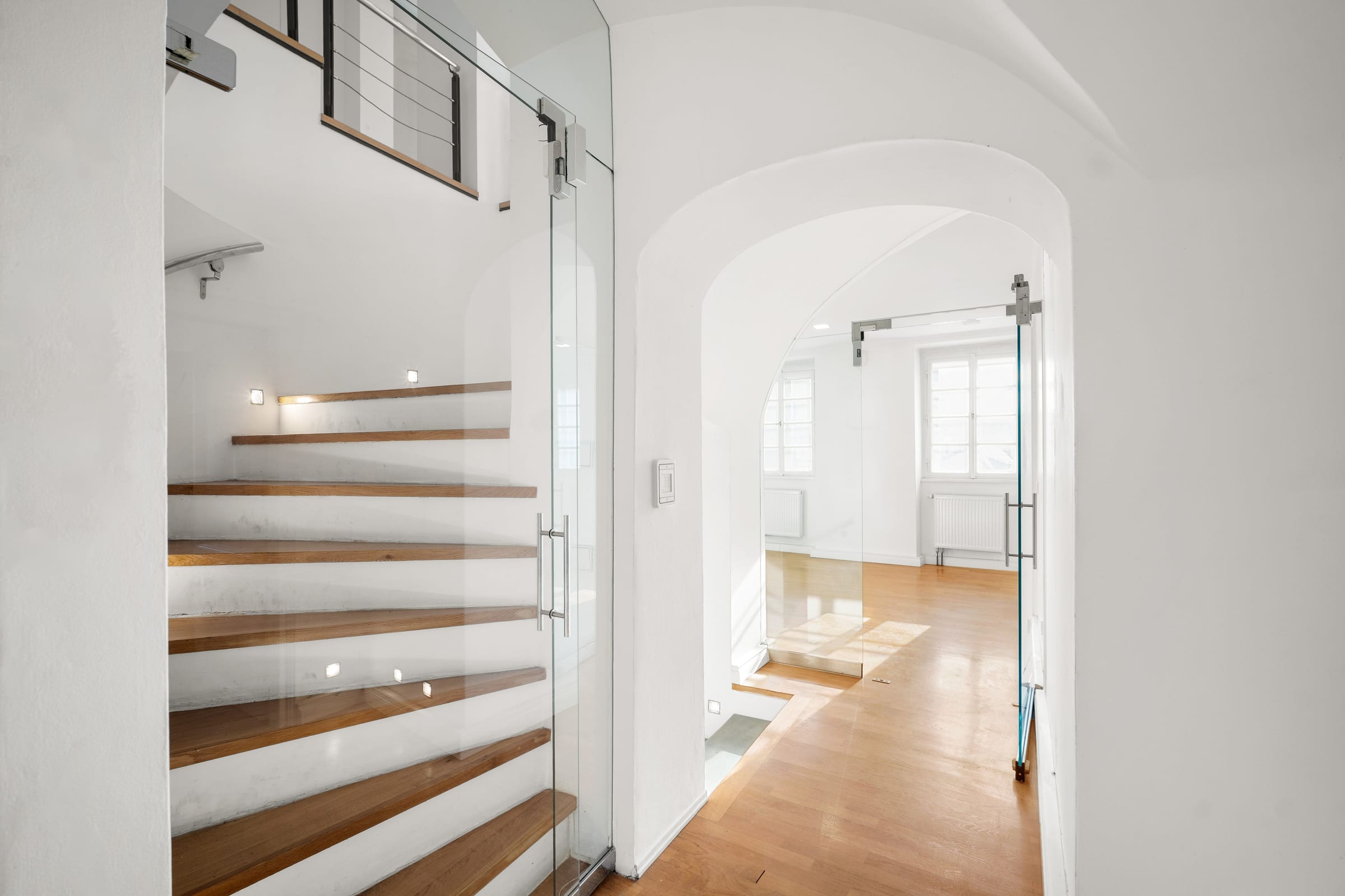 Staircase with wooden treads behind a full-glass door, a vaulted passage beyond
